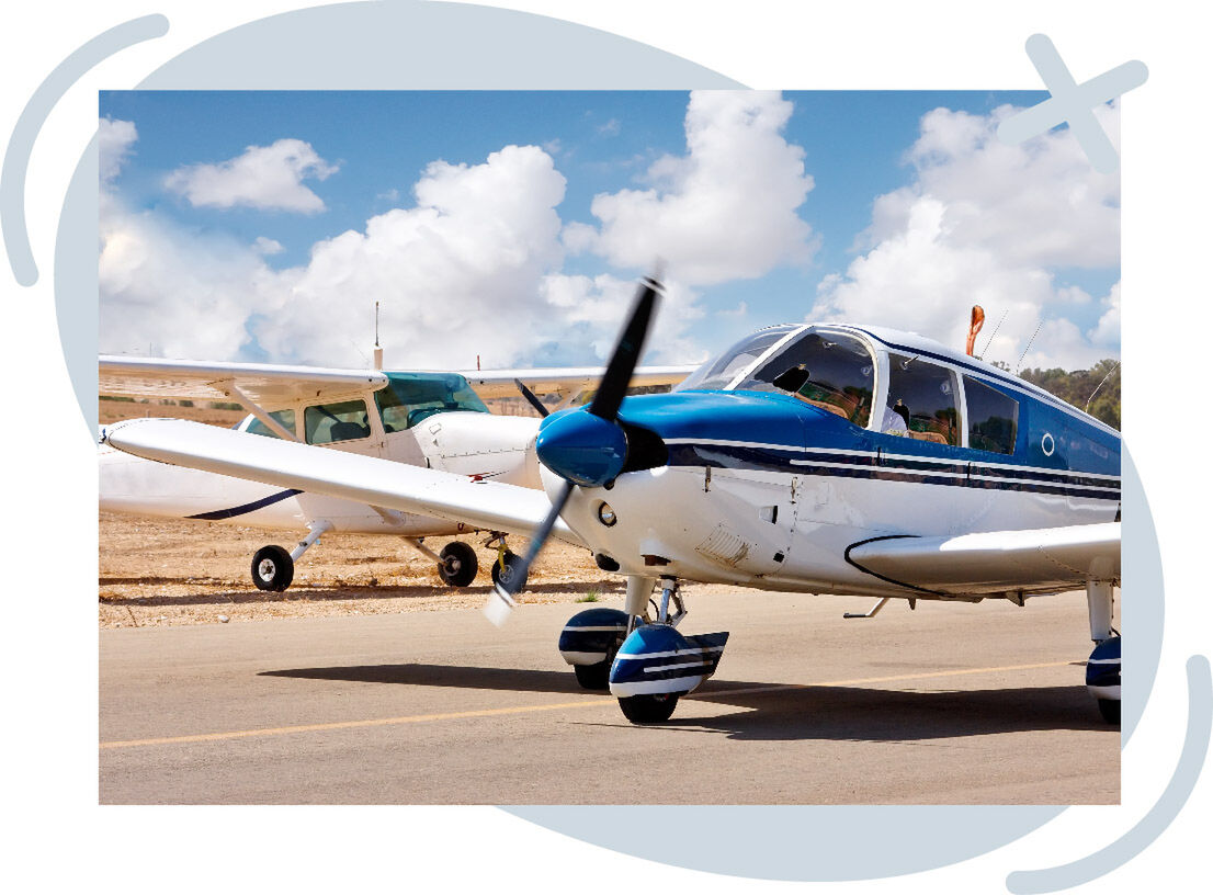 Two small propeller airplanes parked on a sunny airfield, with a blue-and-white aircraft in the foreground and a white plane behind it under a sky of fluffy clouds.