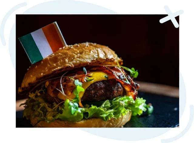 Close-up of a gourmet burger on a dark background, topped with melted cheese, crispy bacon, greens, and an Irish flag toothpick in a shiny brioche bun.