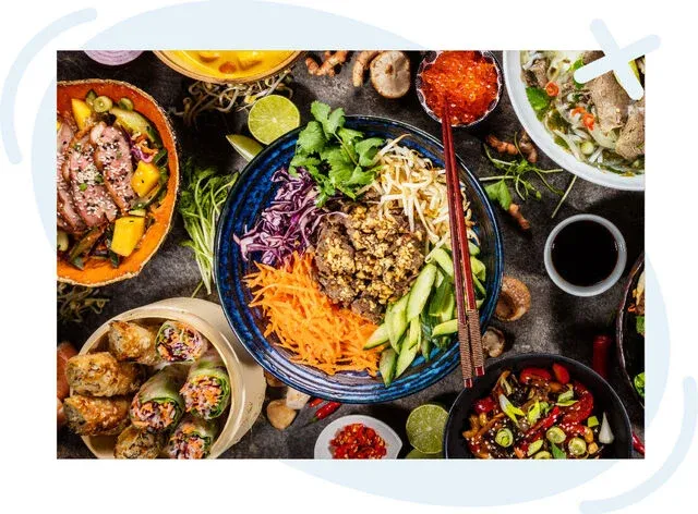 Vibrant overhead spread of Asian dishes with a central noodle bowl topped with colorful vegetables and chopsticks resting on the rim.
