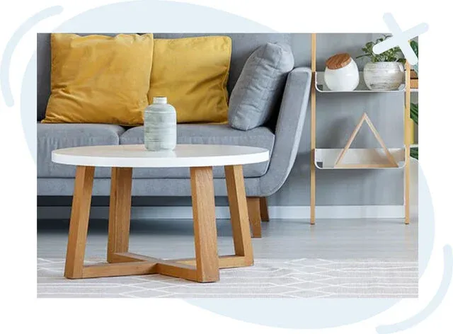 Modern living room scene with a round white coffee table on wooden legs, a light gray sofa with mustard yellow and gray cushions, and a minimalist shelf holding decorative ceramics and a small plant.