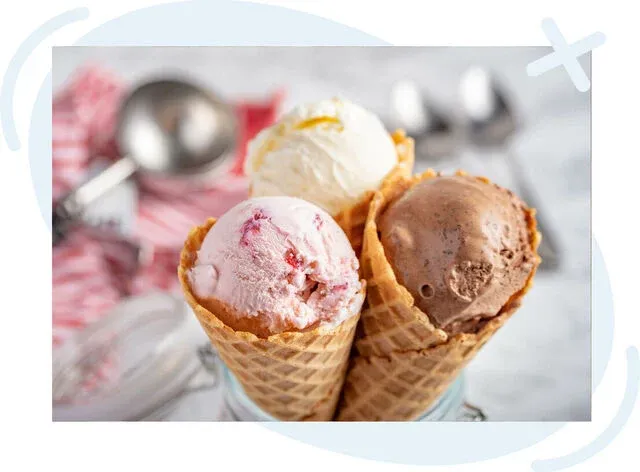 Three waffle cone ice creams arranged in a jar: strawberry with fruit swirls, vanilla with a lemon zest streak, and chocolate with a creamy, slightly textured surface. Soft, bright lighting highlights the scoops, with blurred scoops and striped cloth in t