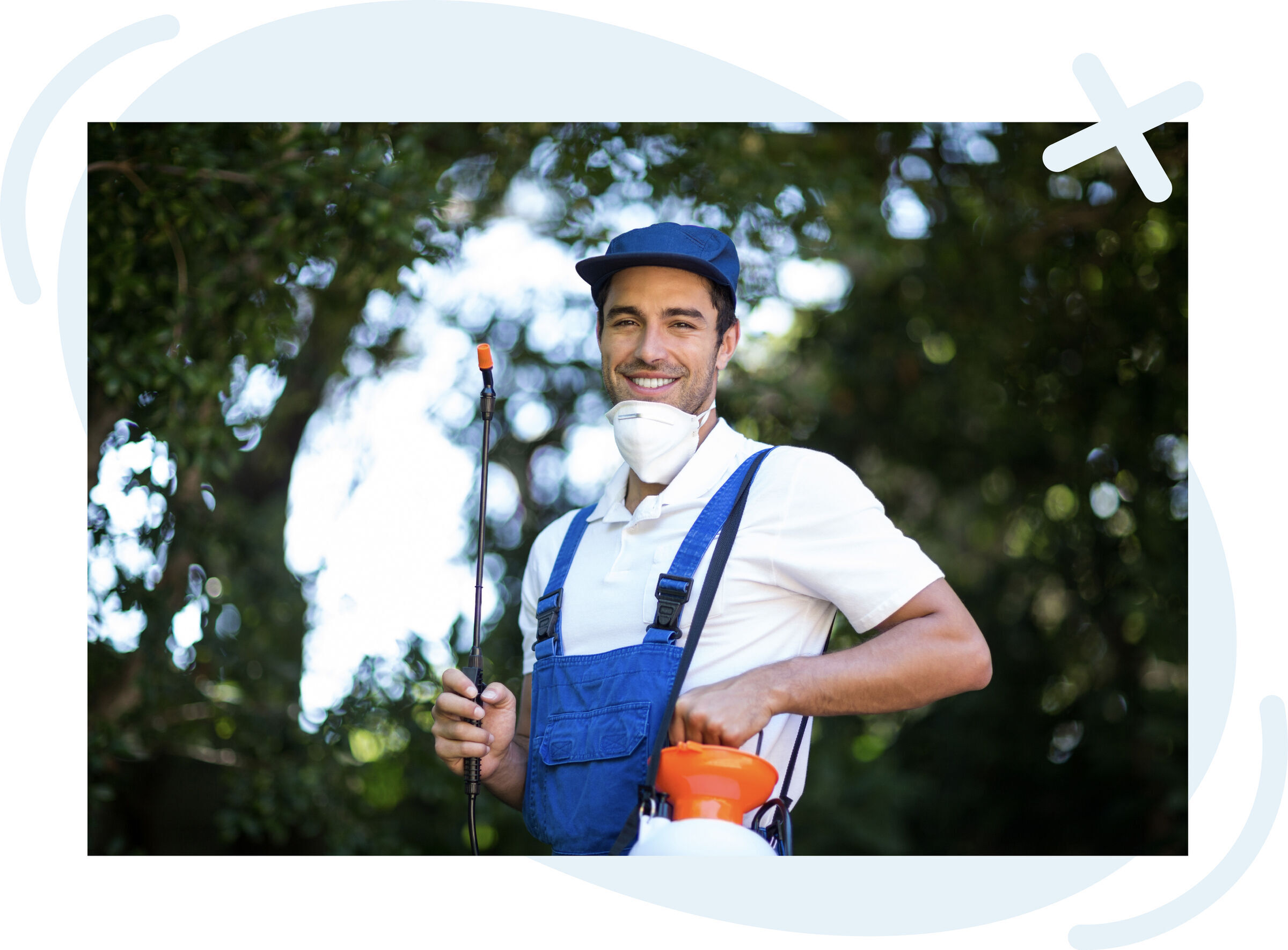 Smiling outdoor worker in blue overalls and cap holding a pesticide sprayer wand with a respirator mask around his neck, standing in a leafy garden.