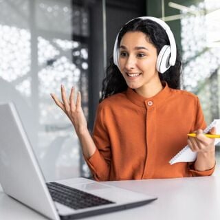 Person wearing white over-ear headphones smiling and gesturing while on a video call at a laptop, holding a notepad and yellow pencil in a modern office.