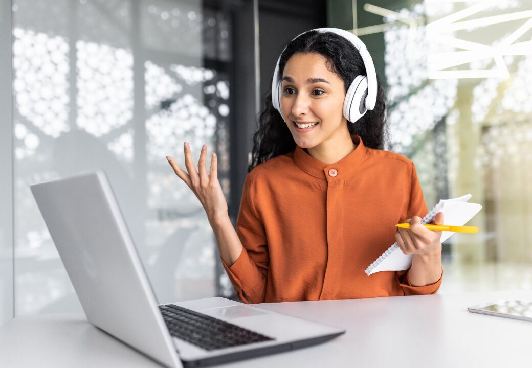 Person wearing white over-ear headphones smiling and gesturing while on a video call at a laptop, holding a notepad and yellow pencil in a modern office.