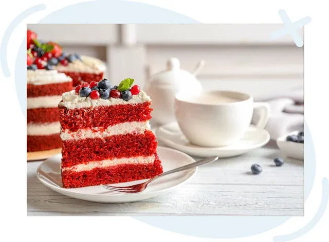 Slice of red velvet layer cake with cream frosting and fresh berries on a white plate beside a white teacup and teapot.