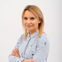 Portrait of a woman with blonde hair standing against a plain white background, arms crossed, wearing a light patterned button-up shirt, looking at the camera with a slight smile.