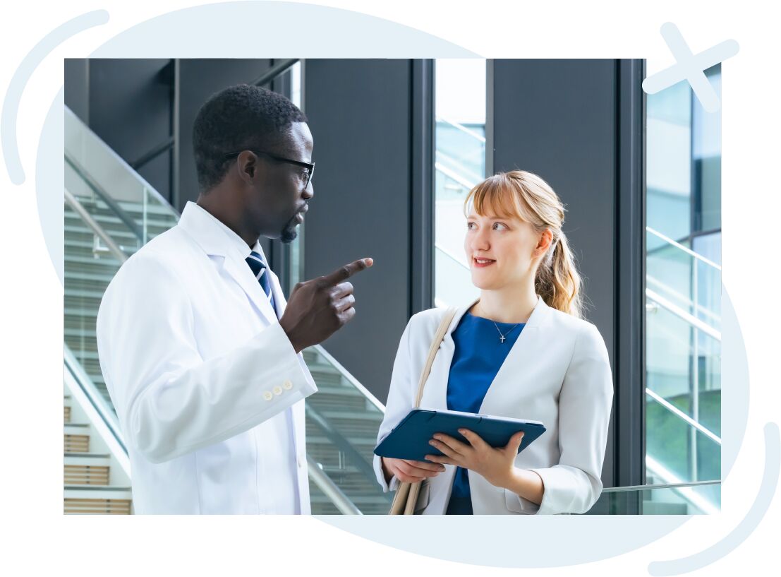 Two professionals in a modern building discuss something; a man in a white lab coat gestures while a woman holding a tablet listens attentively.