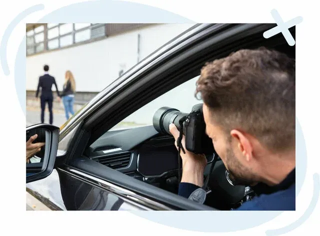 Man inside a car taking photos with a telephoto lens of two people walking together on a city sidewalk.