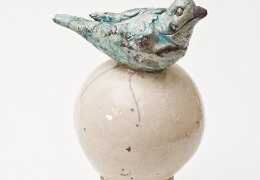 Small bird sculpture with a weathered blue-green patina perched on top of a smooth, round, off-white ceramic sphere against a plain background.