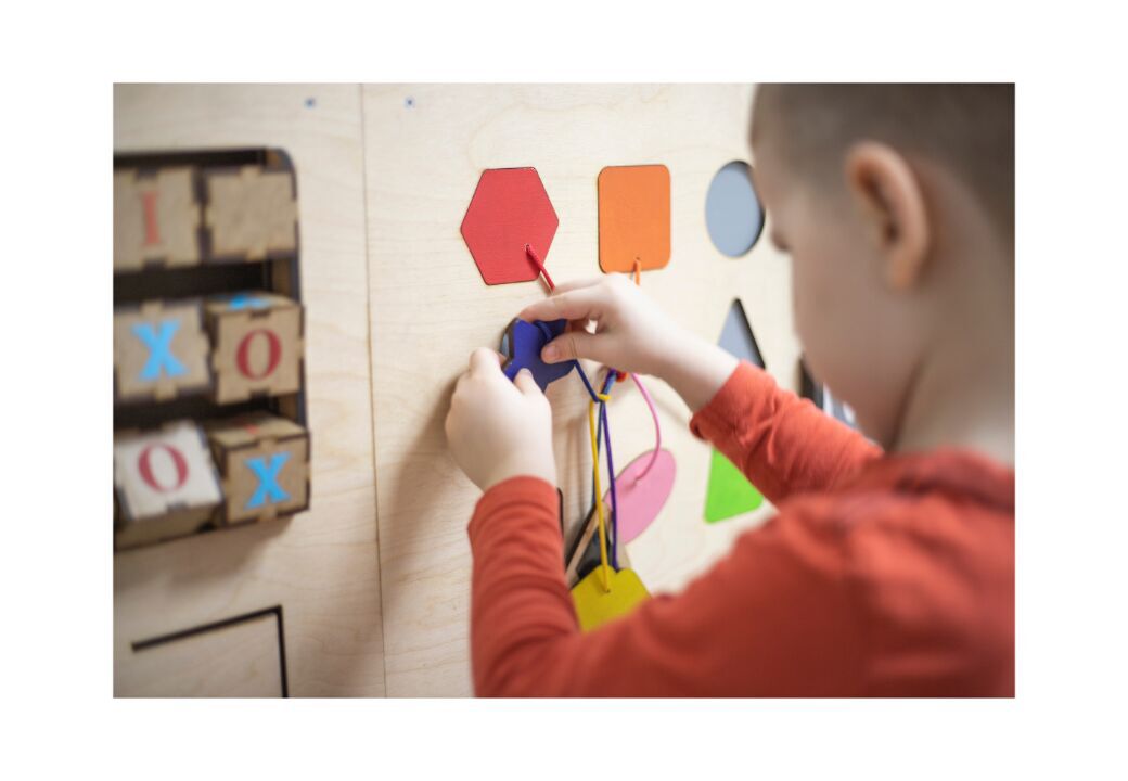 Child playing with a wooden activity board, threading colorful shapes with laces.