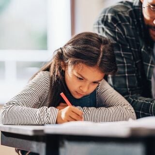 Young student concentrating on writing at a desk with an adult assisting beside her