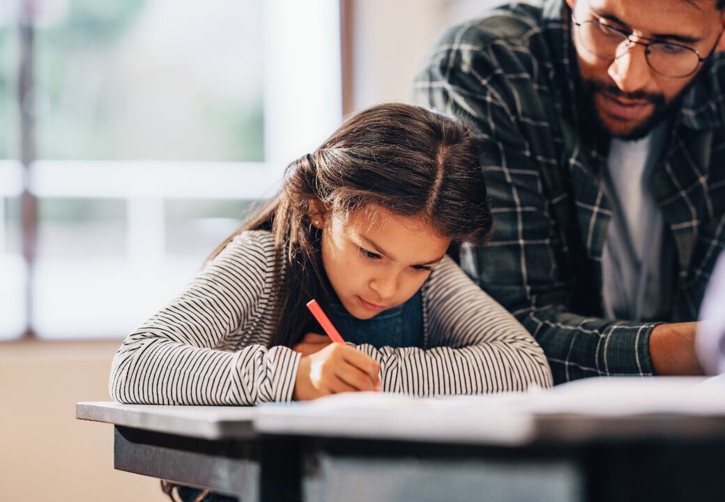 Young student concentrating on writing at a desk with an adult assisting beside her