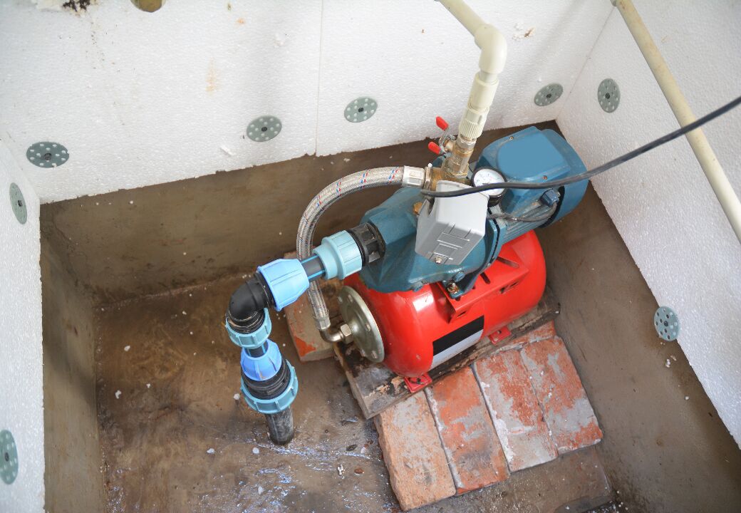 Domestic water pump with red pressure tank installed in a small pit with white insulated walls and brick base.