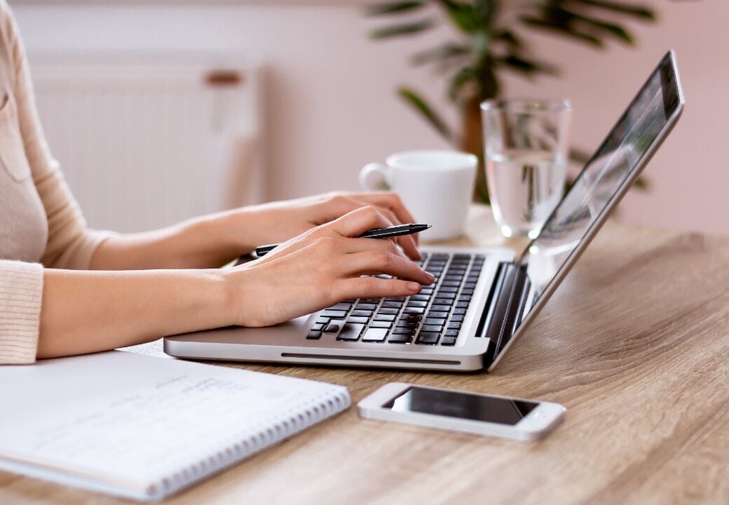 Close-up of a person working on a laptop at a wooden desk with a pen in hand, notebook, smartphone, and a cup and glass of water nearby.