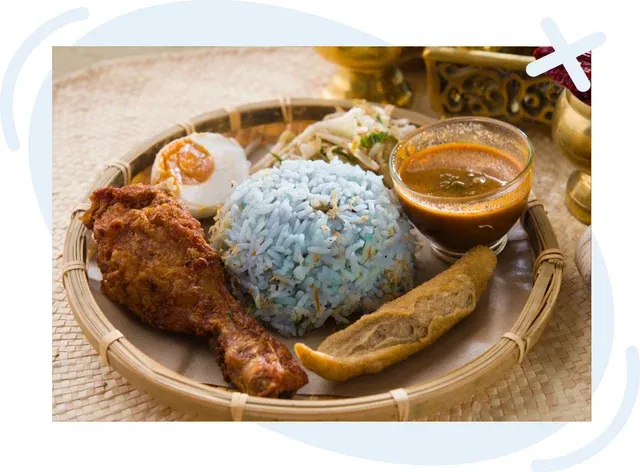 A woven bamboo tray holding a Malaysian-style meal: a mound of light blue-tinted rice in the center, accompanied by a crispy fried chicken drumstick, half of a hard-boiled egg with sambal, a piece of fried fish or tofu, a small bowl of reddish-brown curry