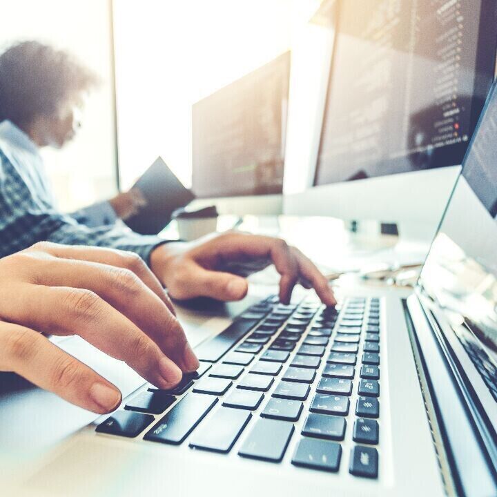 Close-up of hands typing on a laptop keyboard in a bright office with computer monitors displaying code in the background.