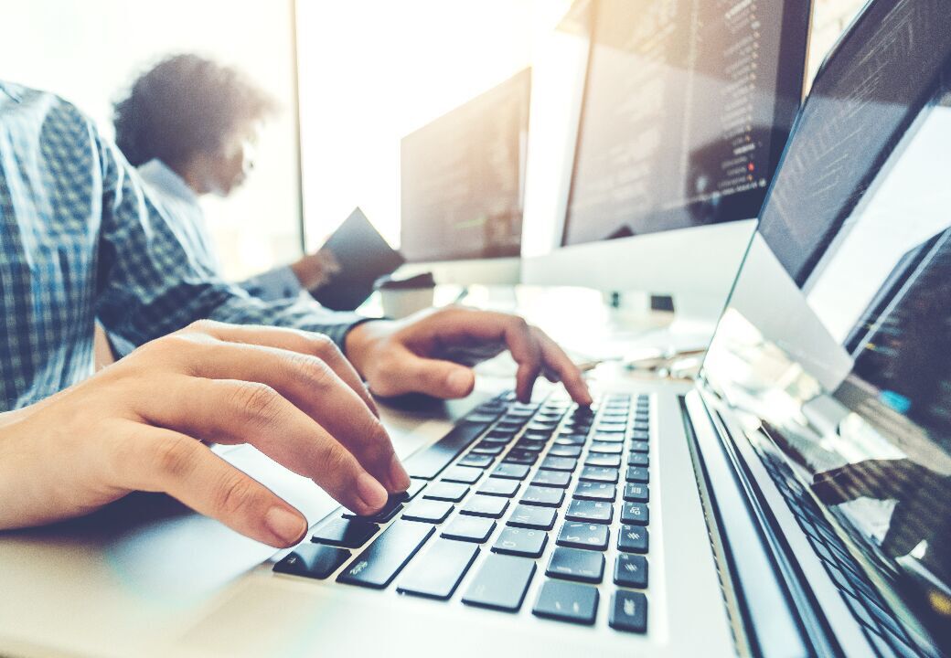 Close-up of hands typing on a laptop keyboard in a bright office with computer monitors displaying code in the background.