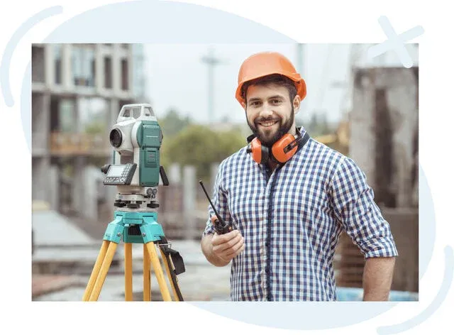 Construction surveyor wearing an orange hard hat and checkered shirt stands smiling at a building site beside a green total station on a yellow tripod, holding a walkie‑talkie with hearing protection resting around his neck.