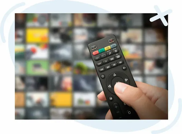 Hand holding a TV remote control pointed at a television screen displaying a grid of blurred streaming thumbnails.