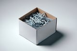 Open cardboard box filled with shiny metal nails on a neutral background.