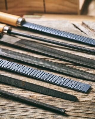 Assorted metal files and rasps with wooden handles laid out on a rustic wooden workbench, showing different shapes and tooth patterns used for shaping wood or metal.