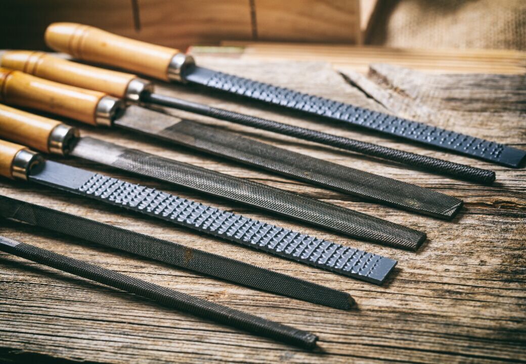 Assorted metal files and rasps with wooden handles laid out on a rustic wooden workbench, showing different shapes and tooth patterns used for shaping wood or metal.
