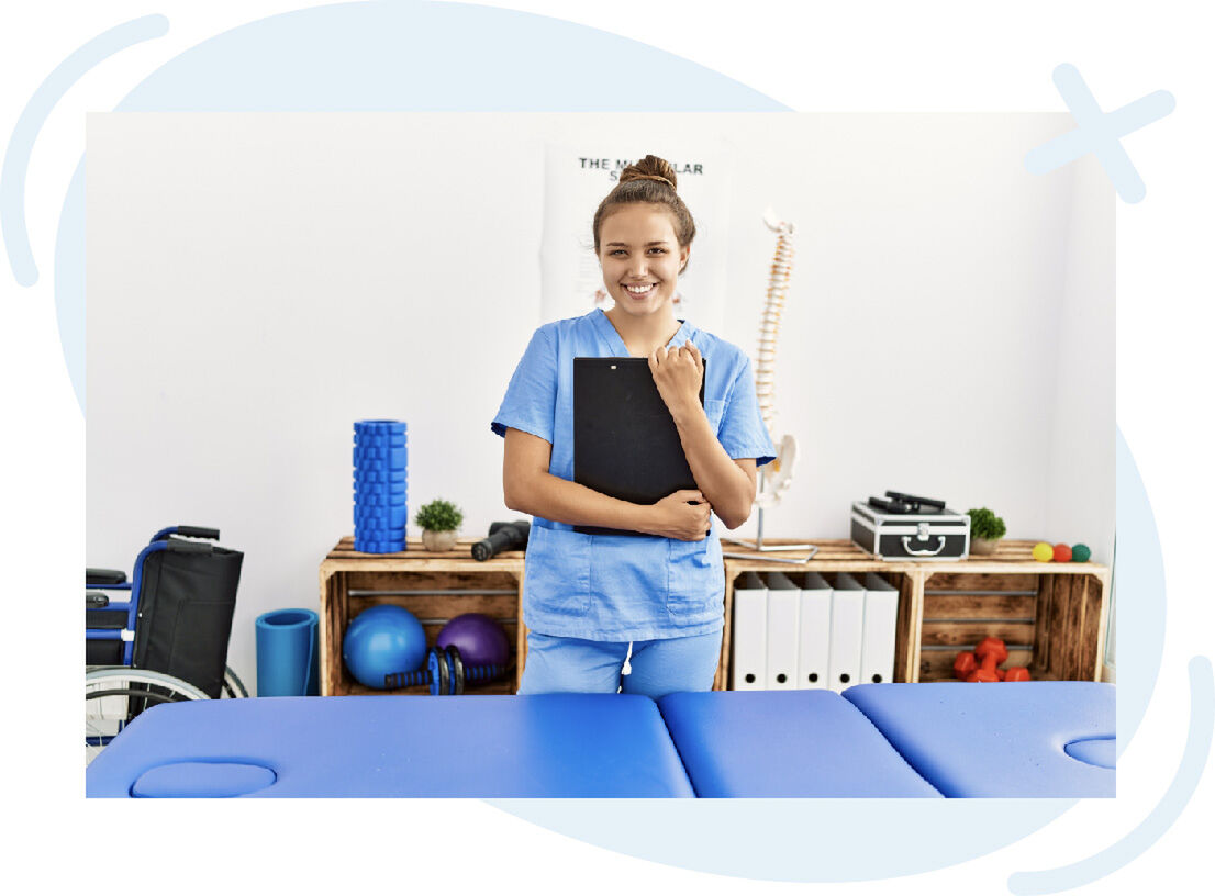 Smiling healthcare professional in blue scrubs holding a clipboard in a therapy room with equipment.