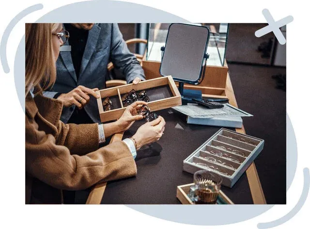 Customer examining eyeglass frames at an optician’s counter with display trays and a small mirror.
