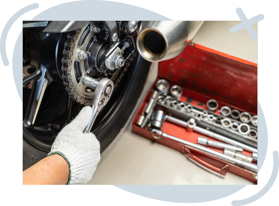 Gloved hand using a ratchet wrench on a motorcycle’s rear sprocket and chain, with an open red toolbox of sockets and tools nearby.