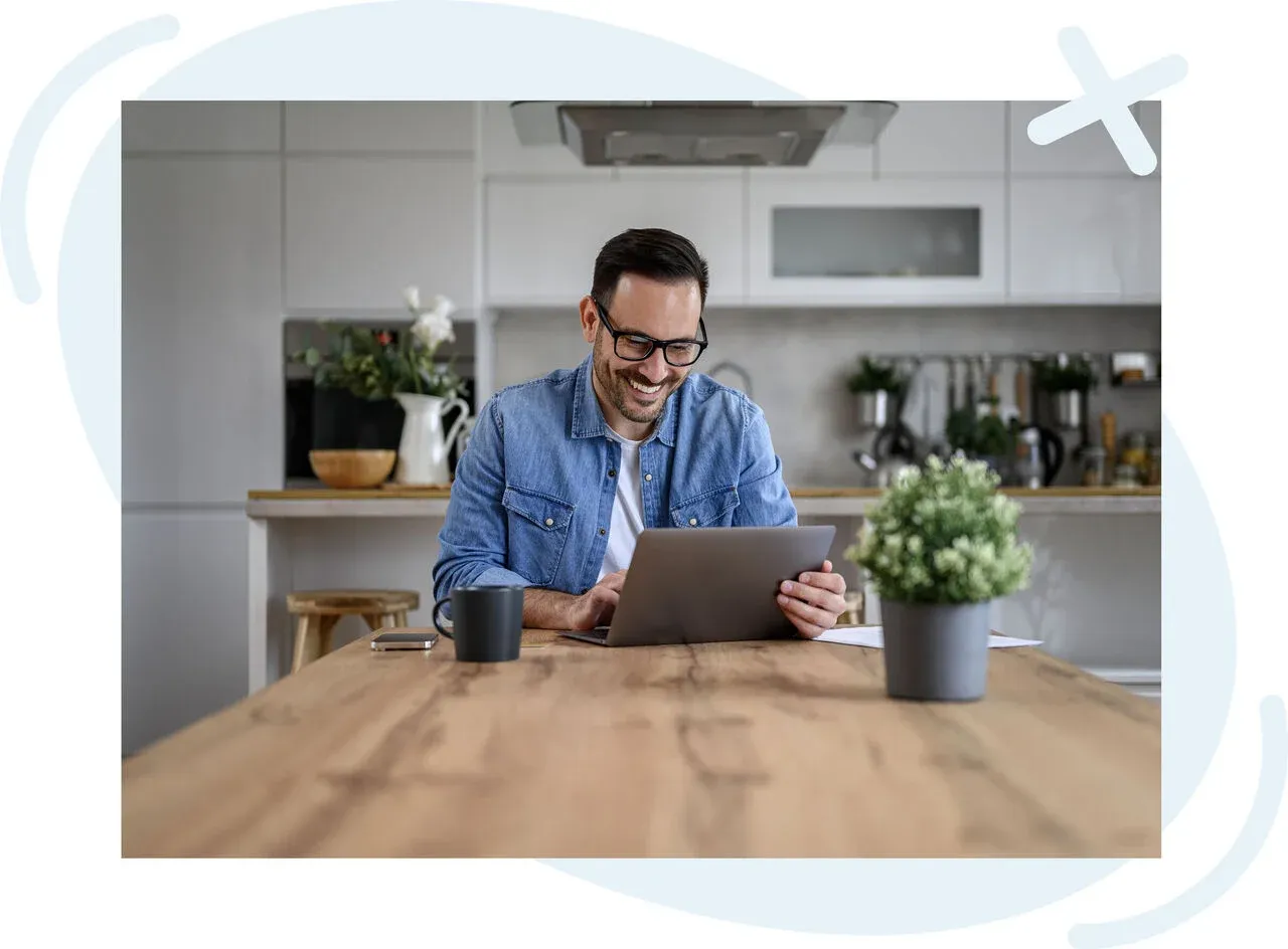 Smiling person using a laptop at a wooden kitchen table in a bright, modern home.