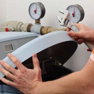 Person servicing a home water softener unit, lifting the lid while holding an adjustable wrench near pipes with pressure gauges.