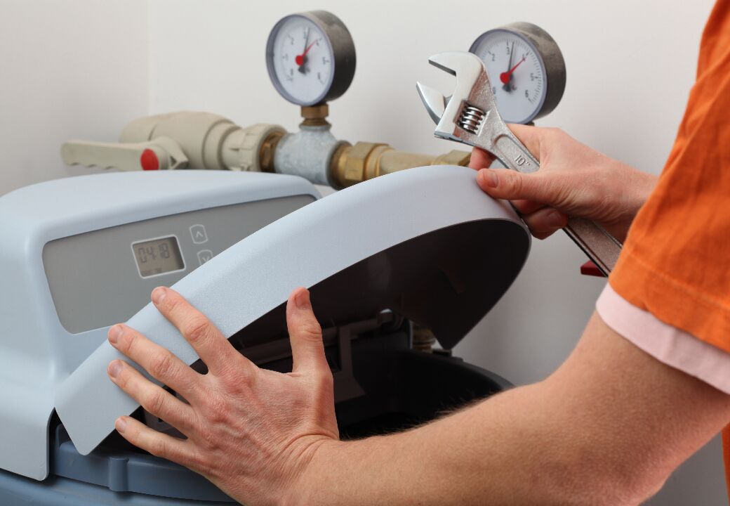 Person servicing a home water softener unit, lifting the lid while holding an adjustable wrench near pipes with pressure gauges.