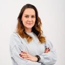 Portrait of a woman standing against a plain white background with arms crossed, wearing a light gray sweater and a smartwatch, looking confidently at the camera.