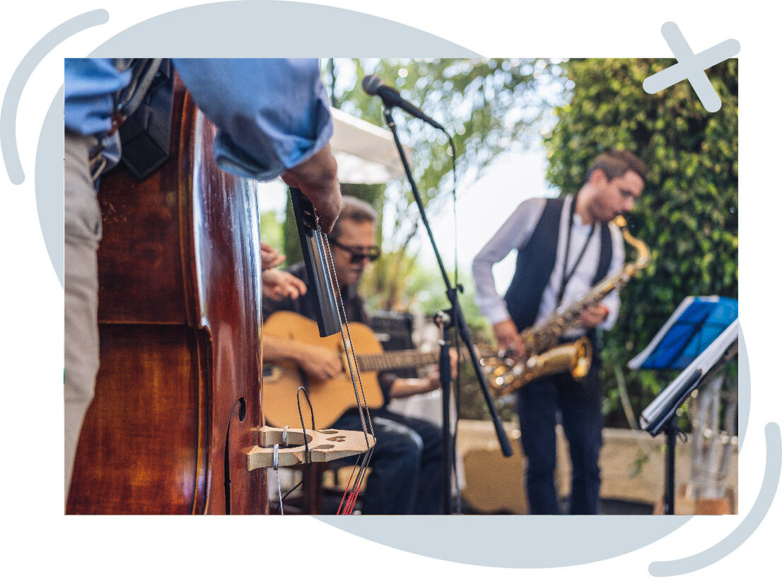 Outdoor jazz band performing with double bass in foreground, guitarist seated, and saxophonist playing near a microphone stand.