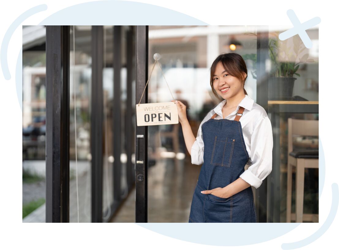 Smiling shop worker in denim apron holding a hanging sign that reads Welcome Open at a glass door entrance.
