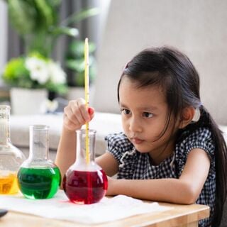 Focused young girl experimenting with colorful liquids in glass flasks at a small table indoors