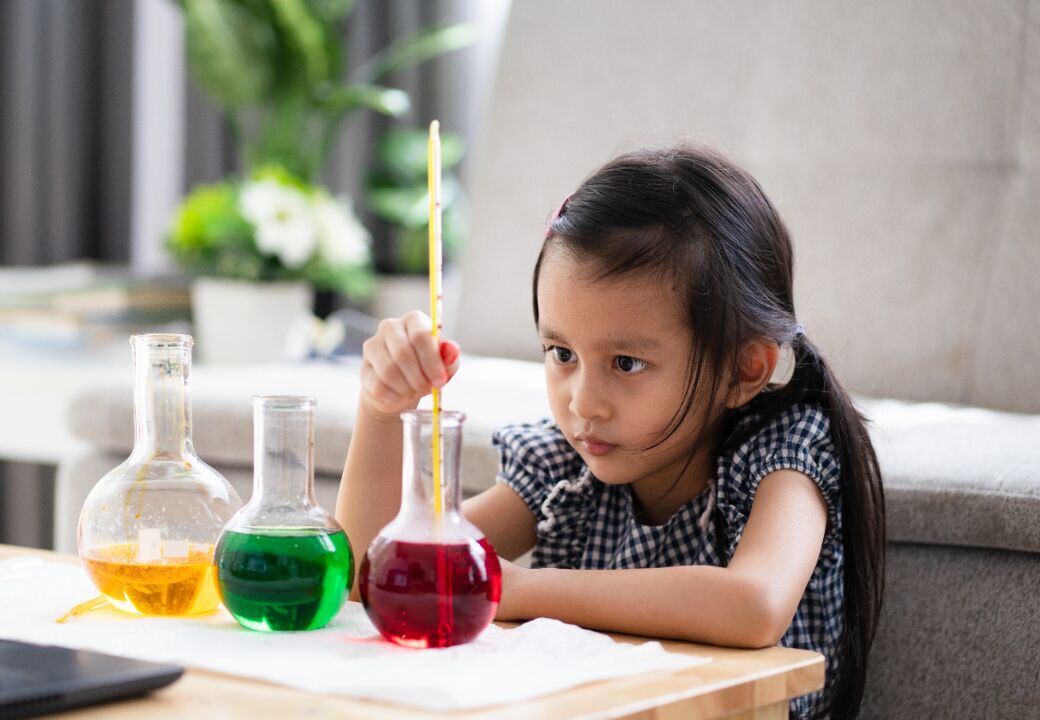 Focused young girl experimenting with colorful liquids in glass flasks at a small table indoors