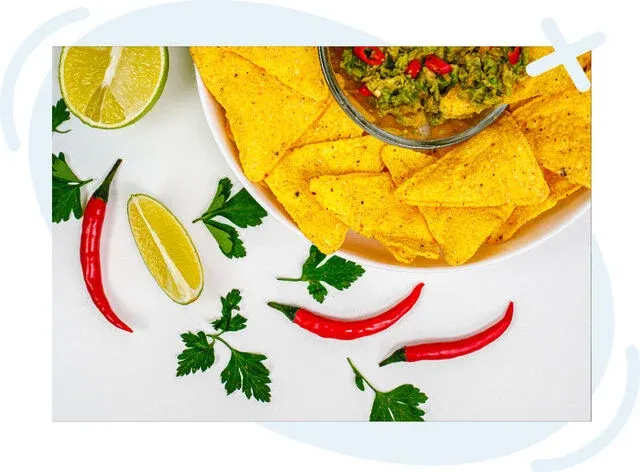 Top-down view of a Tex-Mex snack spread with a bowl of yellow corn tortilla chips and guacamole, surrounded by red chili peppers, lime wedges, and sprigs of parsley on a white surface.