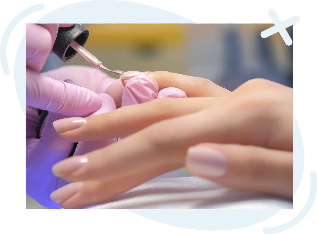 Close-up of a manicurist applying pale pink nail polish to a client’s fingernail with a brush, wearing light pink gloves.