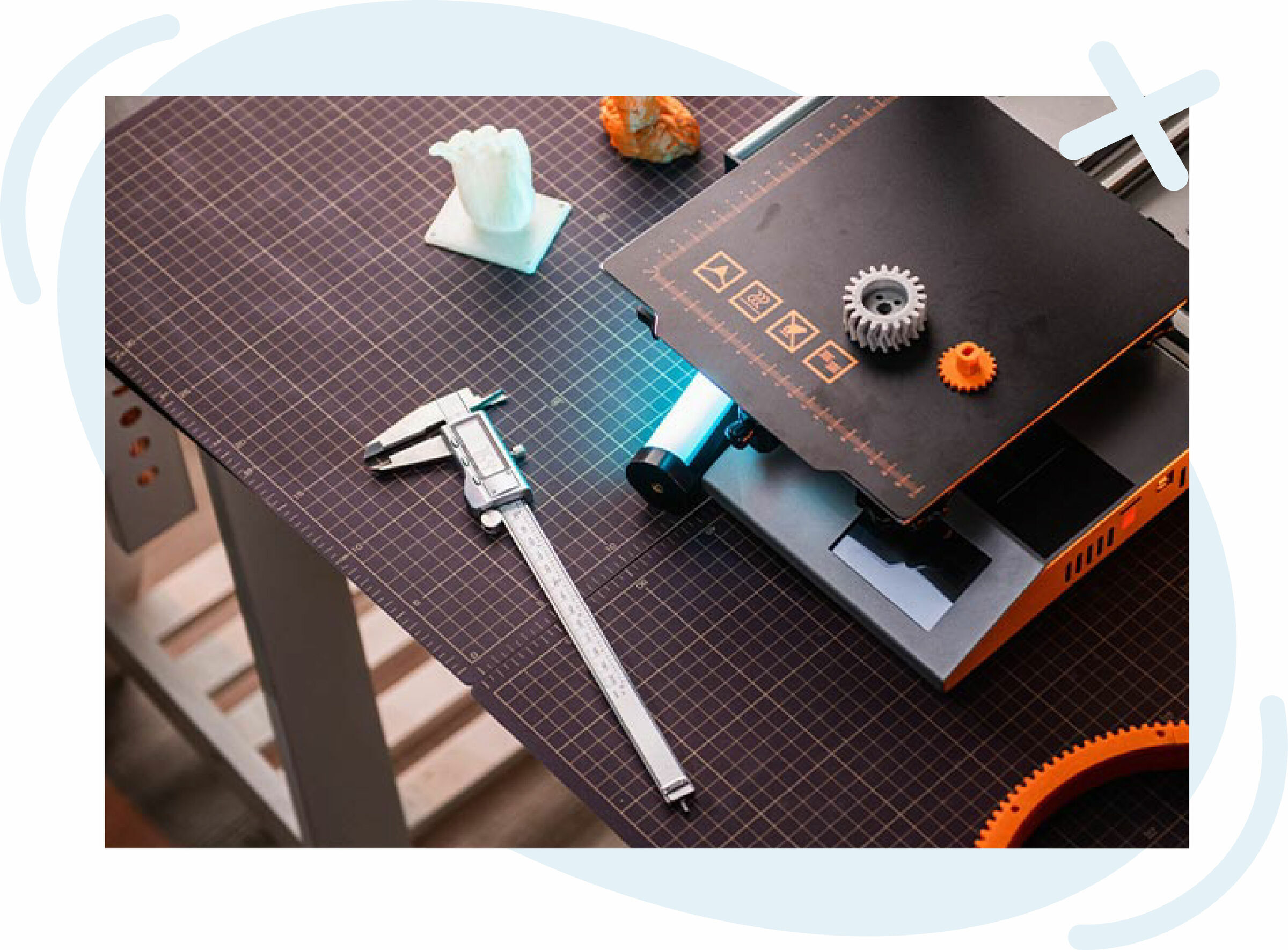 Top view of a 3D printing workspace with a printer, caliper, and printed gears on a gridded cutting mat.