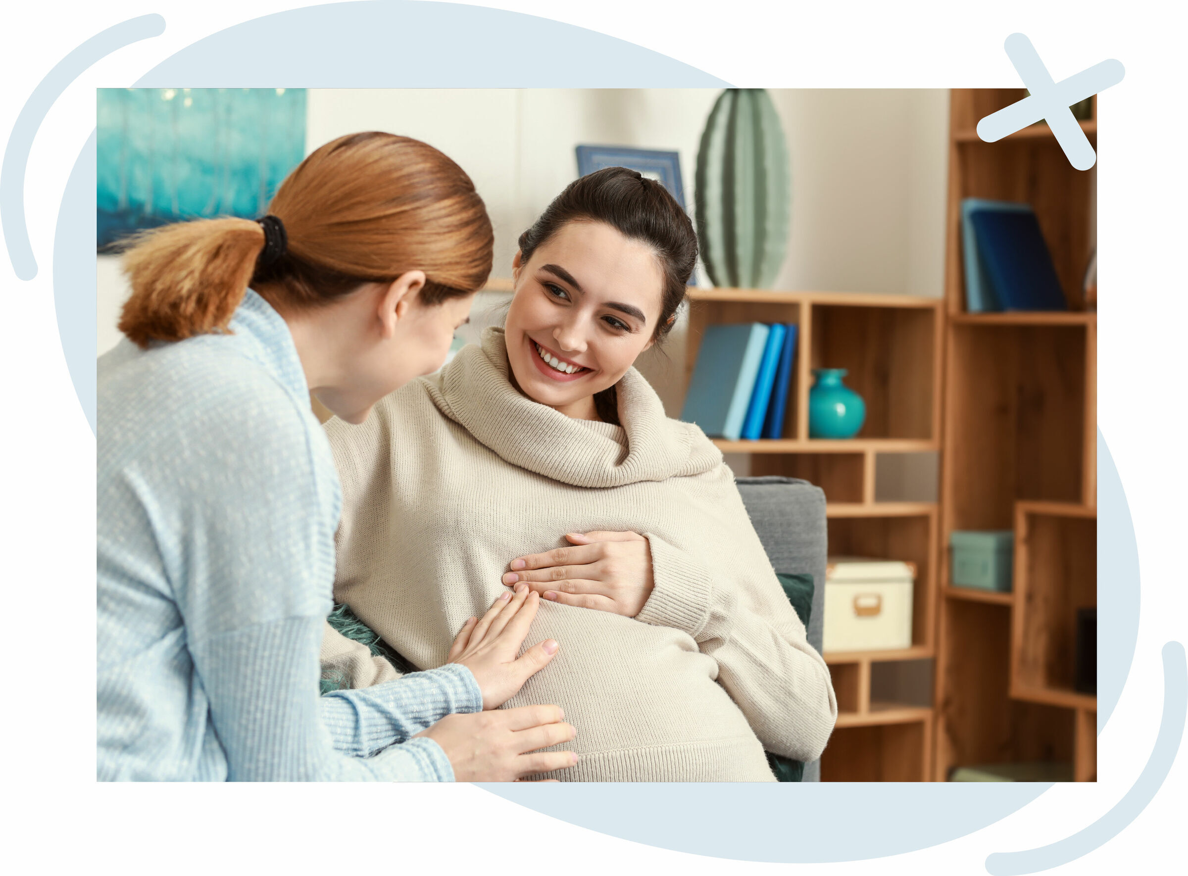 Two women sitting on a couch at home, smiling as they touch a pregnant belly. One woman, wearing a cream turtleneck sweater, is visibly pregnant and looks happily at the other woman in a light blue sweater.