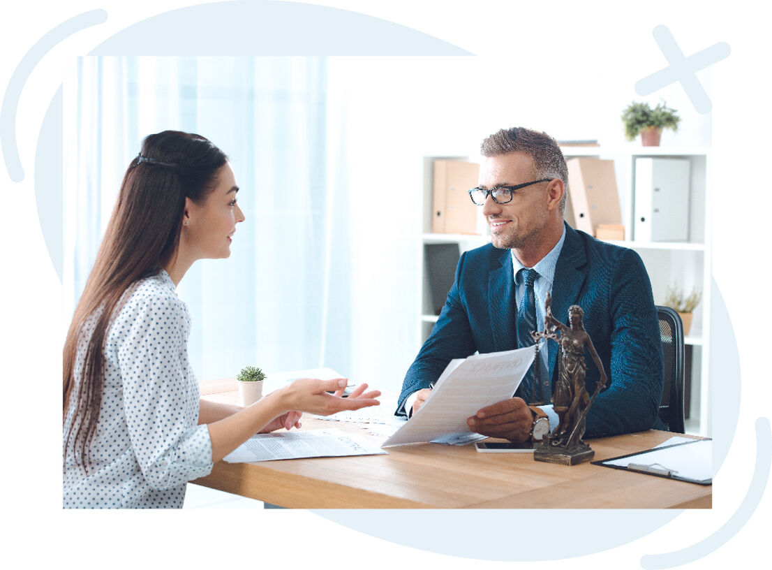 Two professionals talk across a desk in a bright office; a man in a suit reviews documents while a woman gestures as she explains, with a small Lady Justice statue on the desk.
