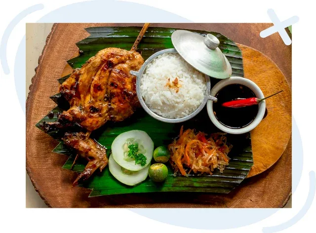Filipino meal on a banana leaf with grilled chicken, steamed rice in a small lidded pot, soy sauce with red chili, and side pickles on a wooden board.