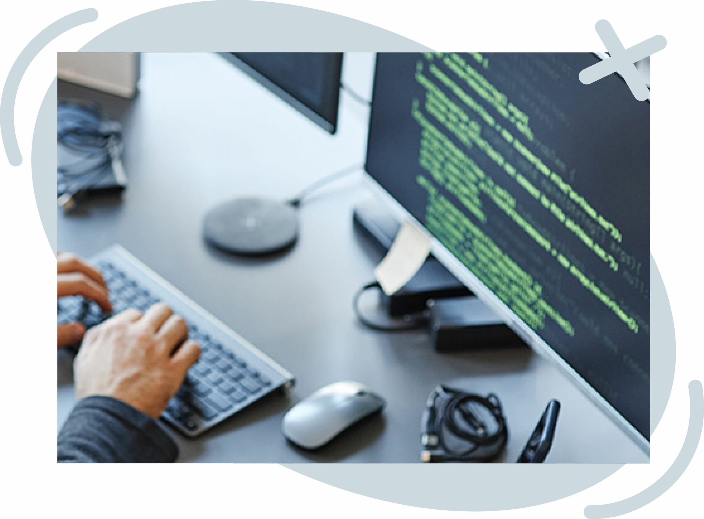 Close-up of hands typing on a keyboard at a desk with a monitor displaying green code on a dark background.