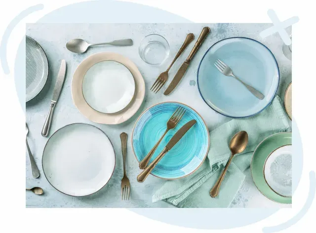 Assorted tableware laid out on a light surface, including colorful plates, cutlery, and glassware in a flat lay arrangement.