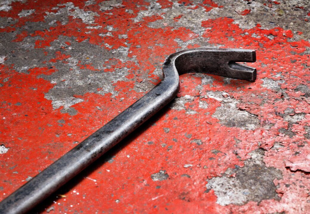 Close-up of a metal pry bar lying on a weathered red concrete surface with peeling paint and gray patches.