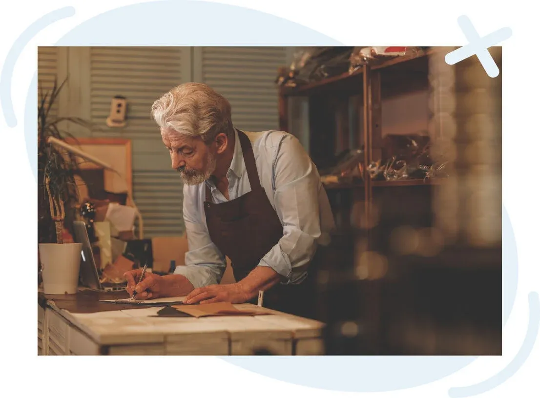 Older man in an apron writing on a clipboard at a workbench in a cozy craft workshop.