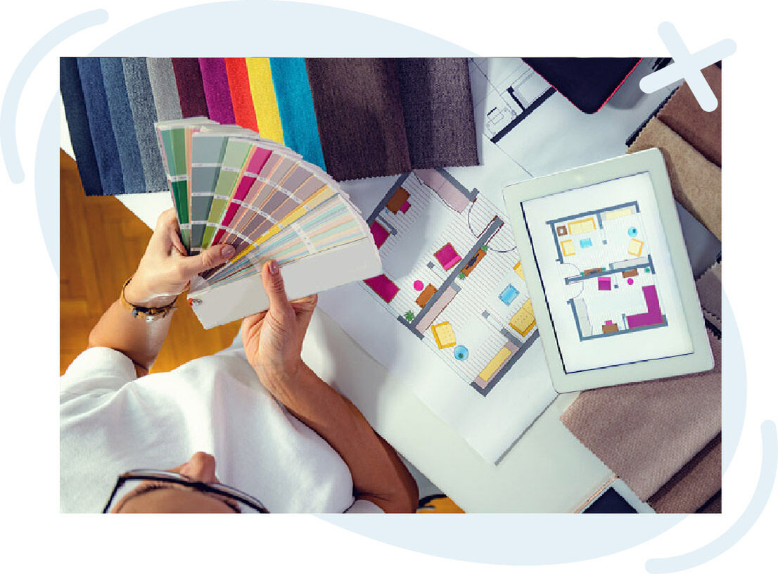Top-down view of a designer selecting paint colors with a swatch fan over floor plans and a tablet showing a room layout, surrounded by fabric samples.