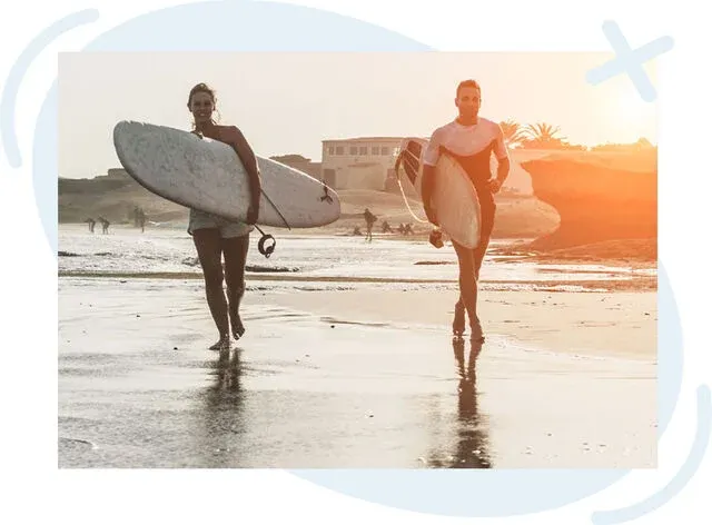 Two surfers carrying boards while walking along a sunlit beach shoreline