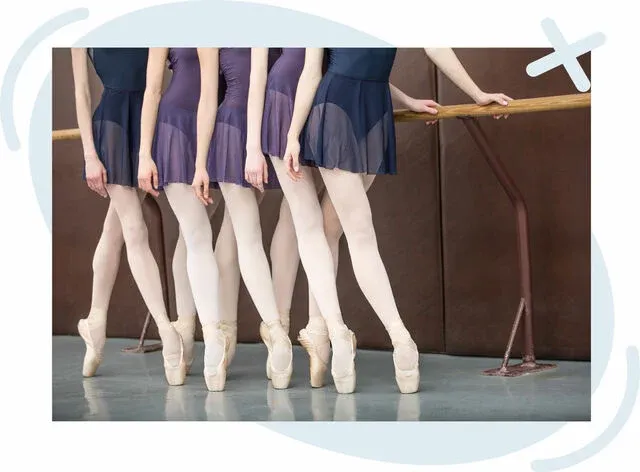 Row of ballet dancers at a studio barre standing en pointe, wearing purple leotards with short skirts, pale tights, and pointe shoes.